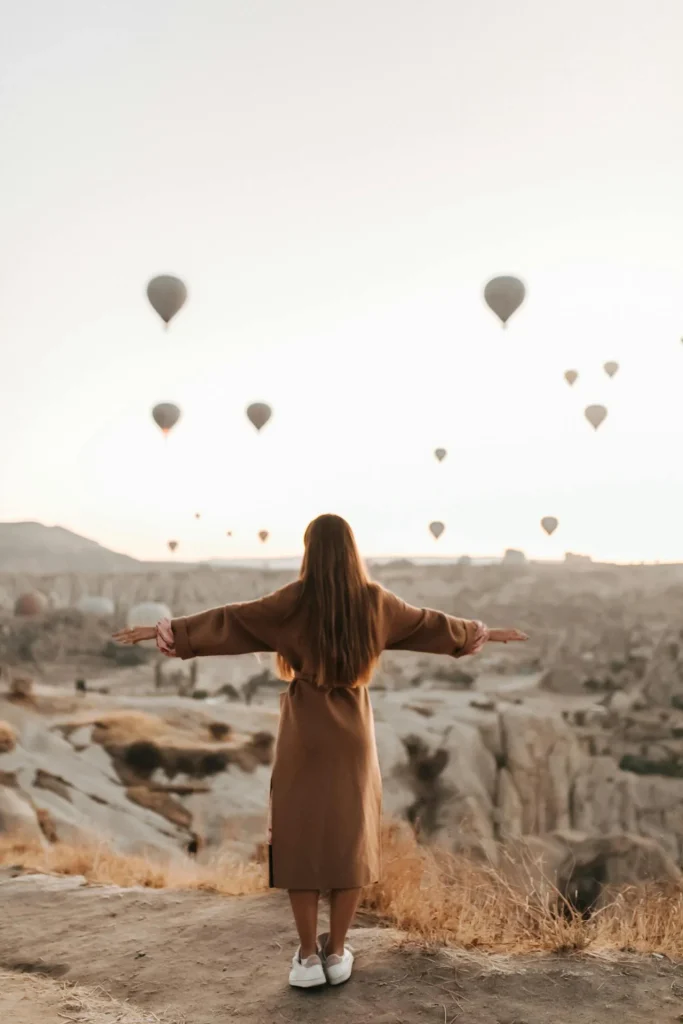 Fairytale Places: a woman with open arms watching hot air balloons over rocky landscapes at sunrise.